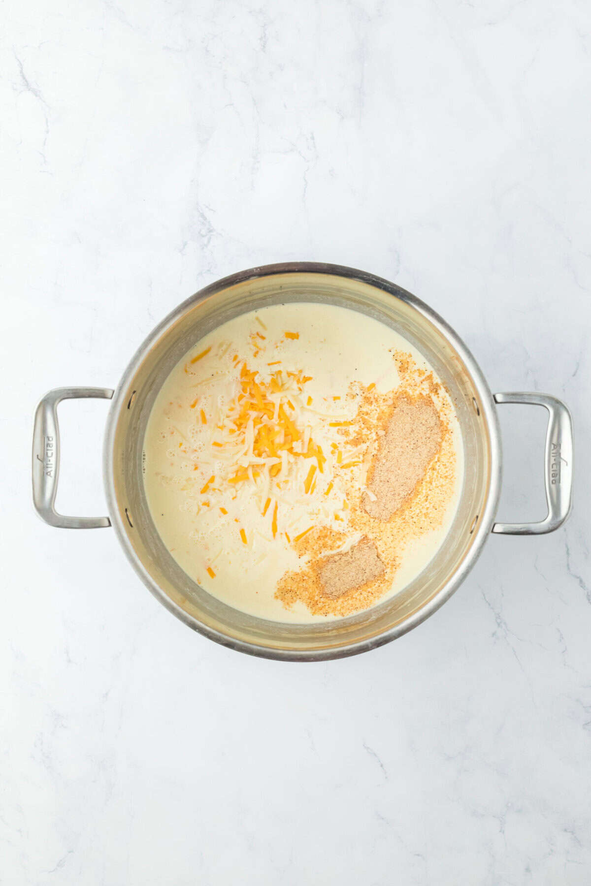 Cream, seasoning and cheese being added to a pot on white countertop.