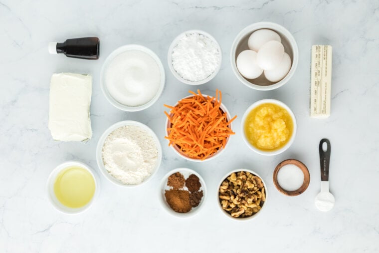 Grated carrots, pineapple, eggs, flour, oil in white bowls on white countertop