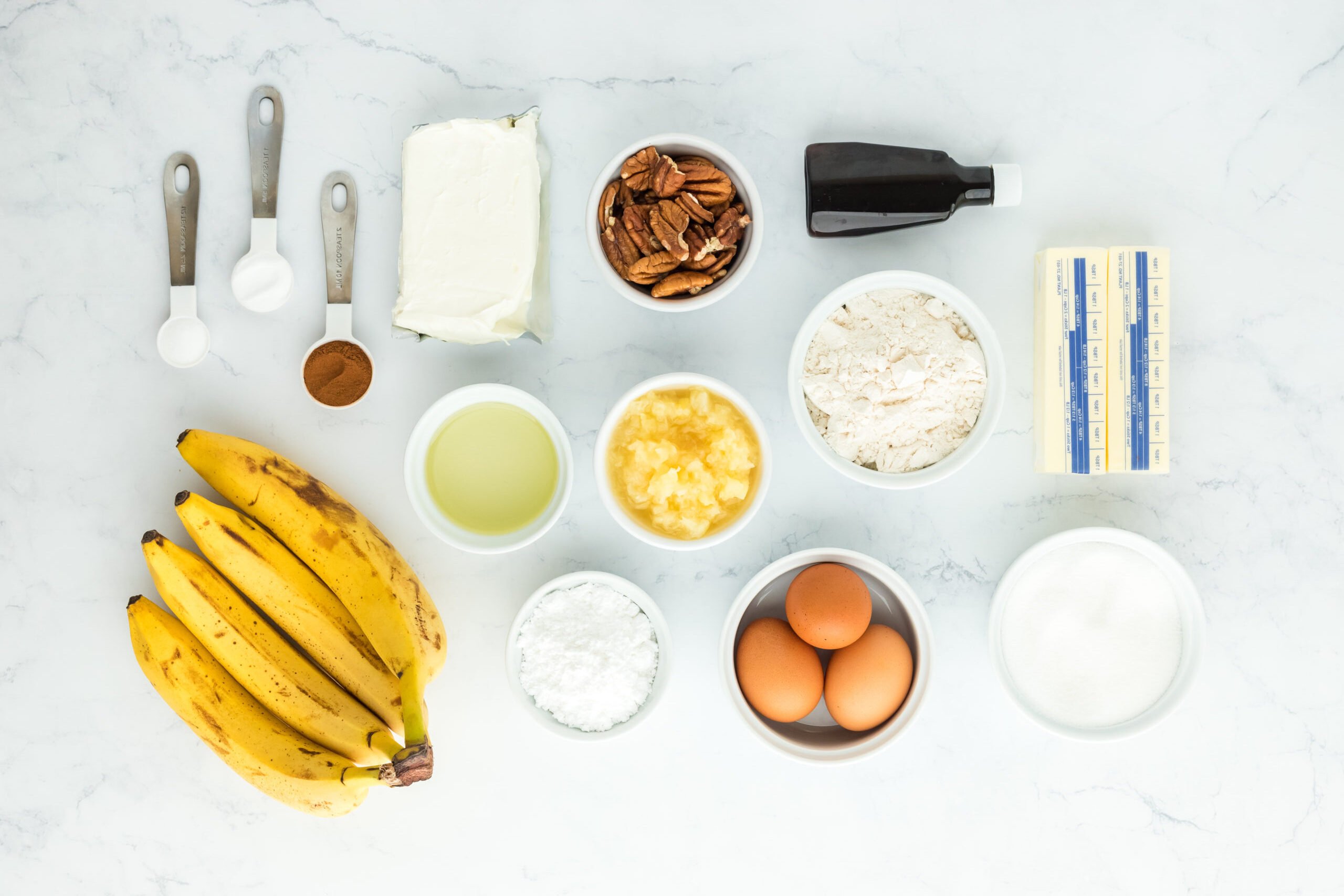 Bananas, eggs, pineapple, and other ingredients in white bowls on white countertop to make a cake