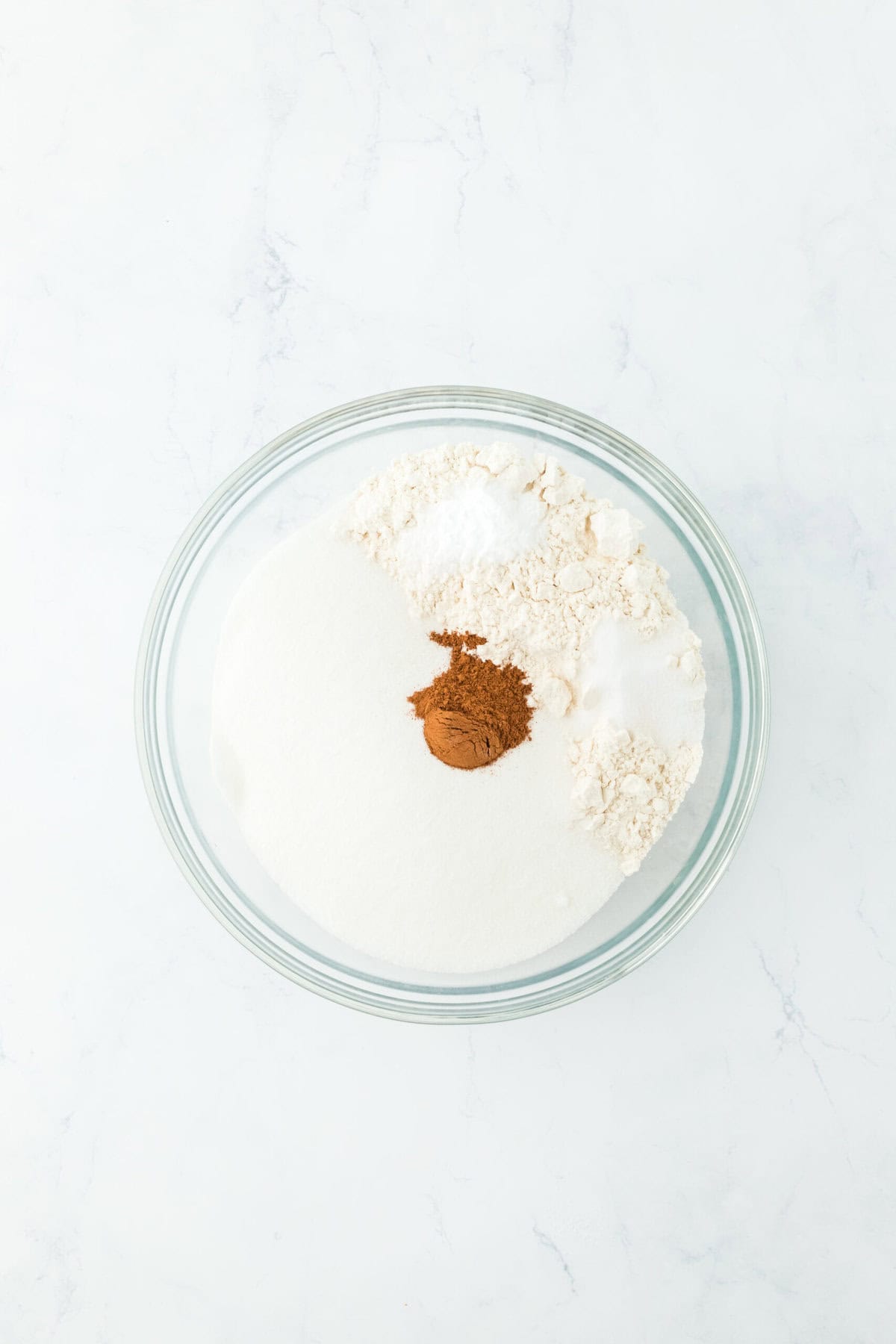 Large glass bowl filled with flour, sugar, cinnamon and other dry ingredients on white countertop