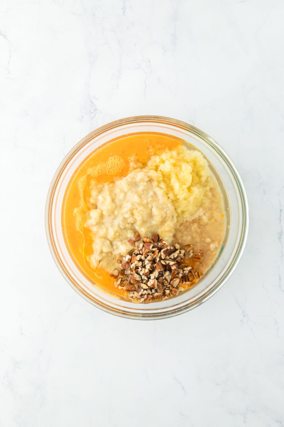 Eggs, oil, vanilla, pineapple, banana and pecans in a glass bowl on white countertop