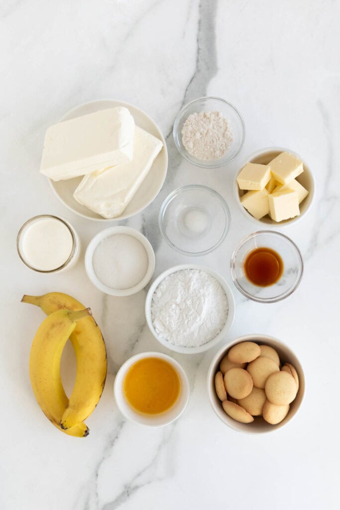 Cream cheese, bananas, heavy cream, butter, nilla wafers and other baking ingredients in glass bowls on white countertop