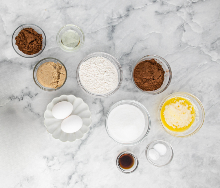 Cocoa, flour, brown sugar, granulated sugar, eggs and vanilla in glass bowls on white countertop