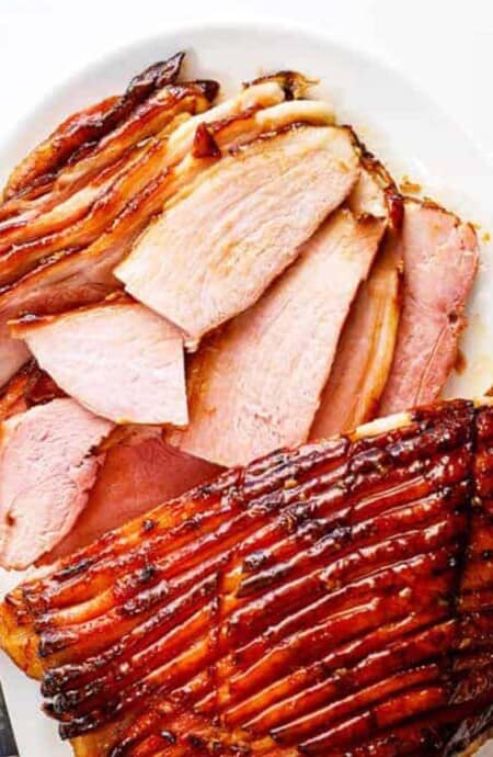 Honey baked ham on a platter with one end sliced up and the other whole with honey glaze.