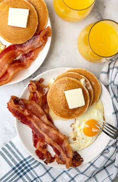 Pancakes piled high with butter, candied bacon and egg along with orange juice being served for breakfast