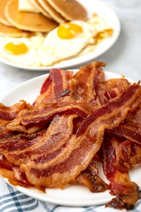 A pile of bacon on a plate with eggs near by for breakfast