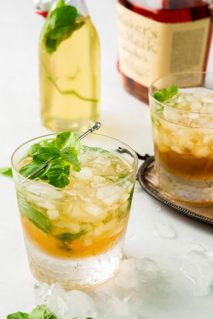 Two mint juleps with bourbon in the background and mint simple syrup