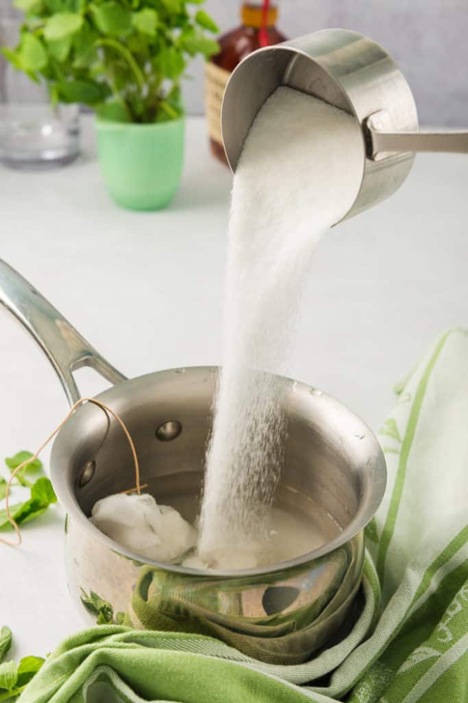 Granulated sugar being poured into water and mint to make a mint simple syrup for cocktails