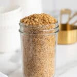 Jar filled with packed homemade brown sugar set on a marble surface with measuring cups behind it
