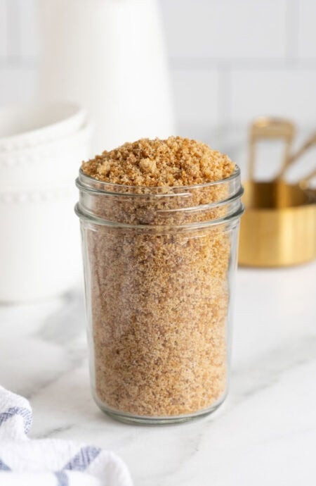 Jar filled with packed homemade brown sugar set on a marble surface with measuring cups behind it
