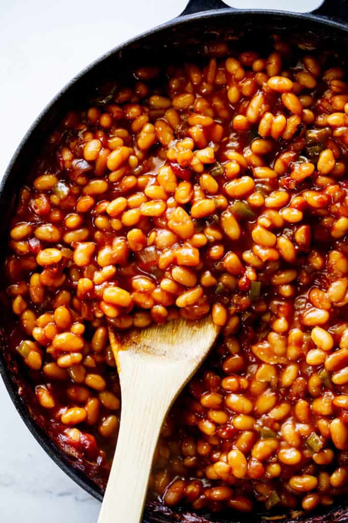 A large wooden spoon serving baked beans in a large skillet