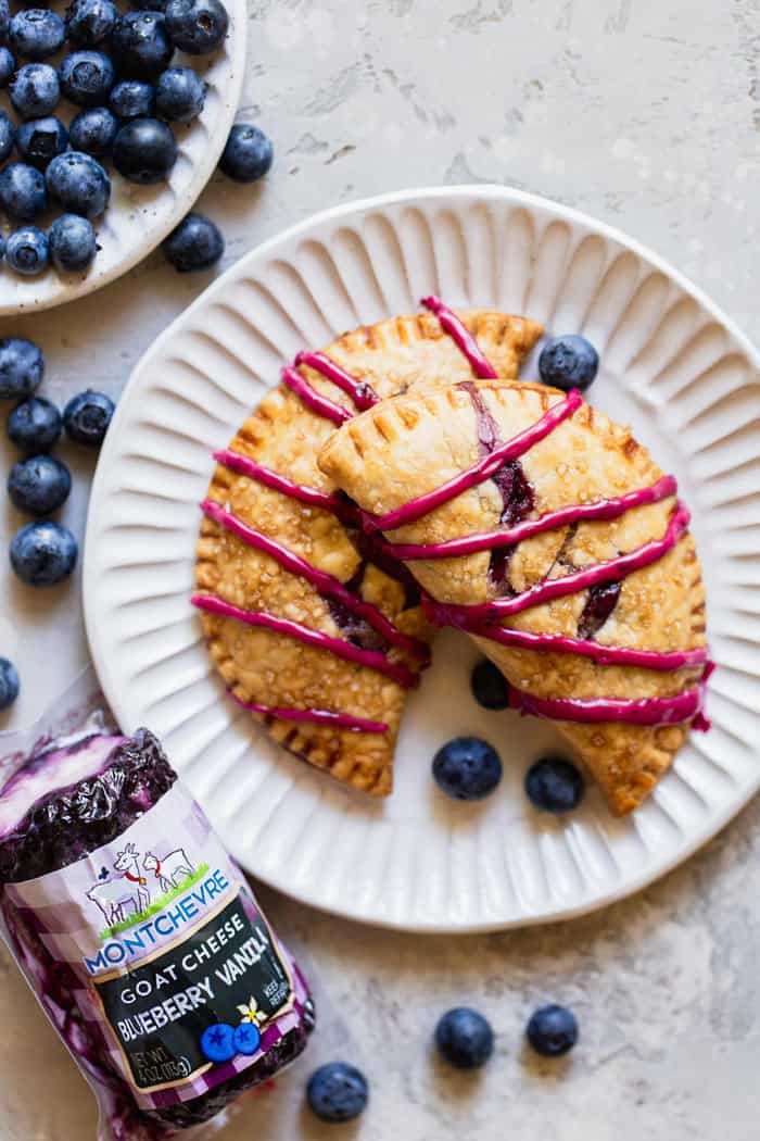 An overhead shot of two blueberry turnovers with blueberry icing ready to serve on a white plate next to fresh blueberries and goat cheese in a package