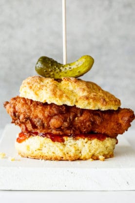 A fried chicken breast on a buttermilk biscuit with red pepper jelly and a whole pickle against a gray background