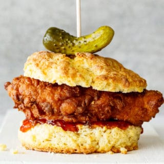 A fried chicken breast on a buttermilk biscuit with red pepper jelly and a whole pickle against a gray background