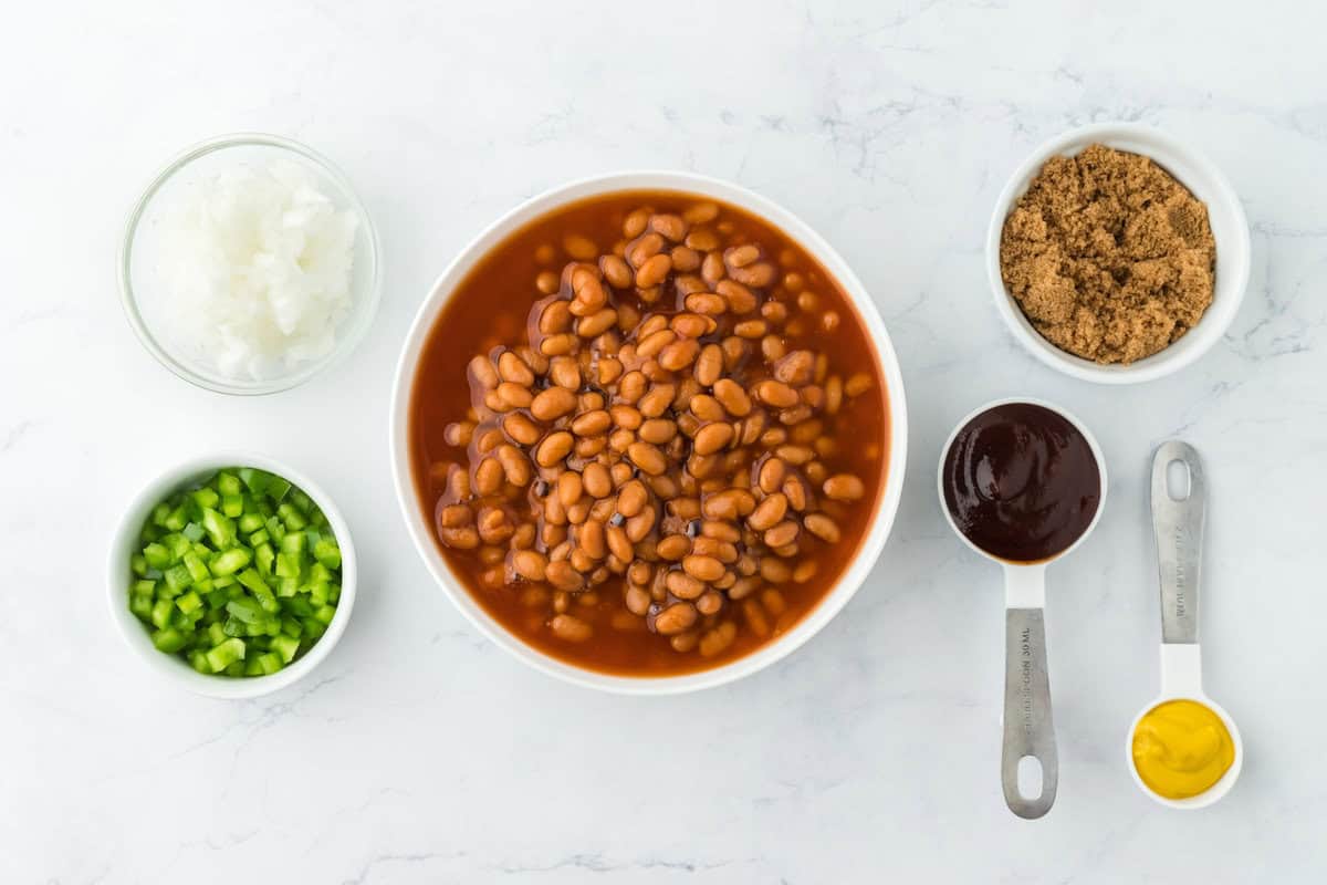 Vegetarian beans, brown sugar, bbq sauce, mustard, and chopped veggies in bowls and measuring spoons on a white background