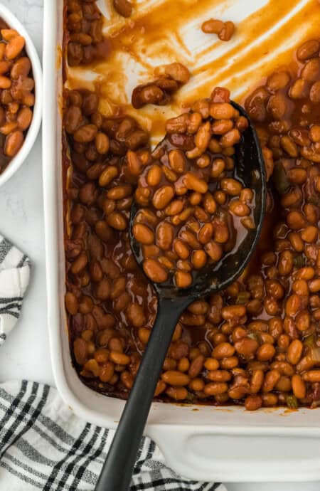 A close up of the best baked beans in a white casserole dish with a spoon scooping some to serve