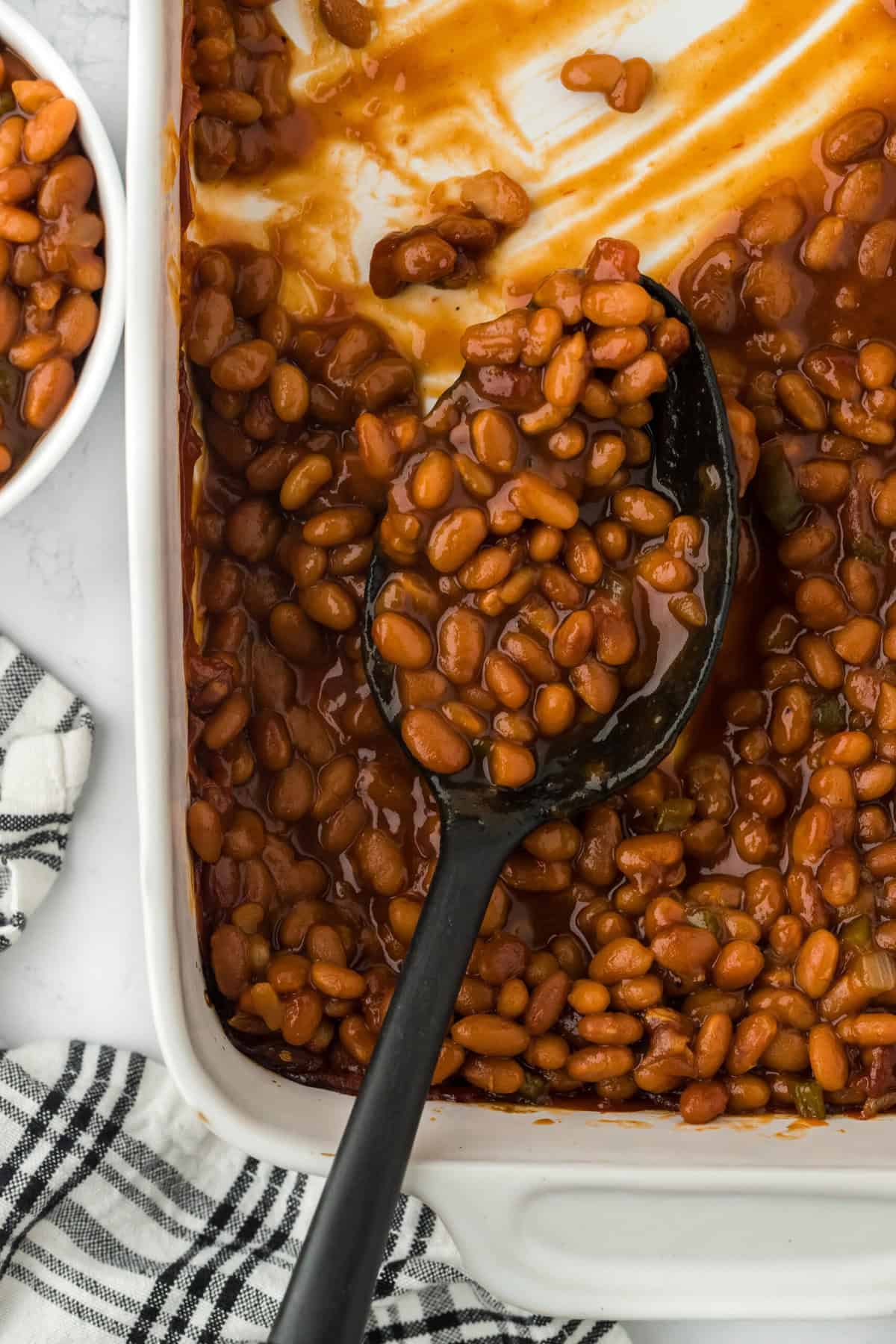 A close up of the best baked beans in a white casserole dish with a spoon scooping some to serve