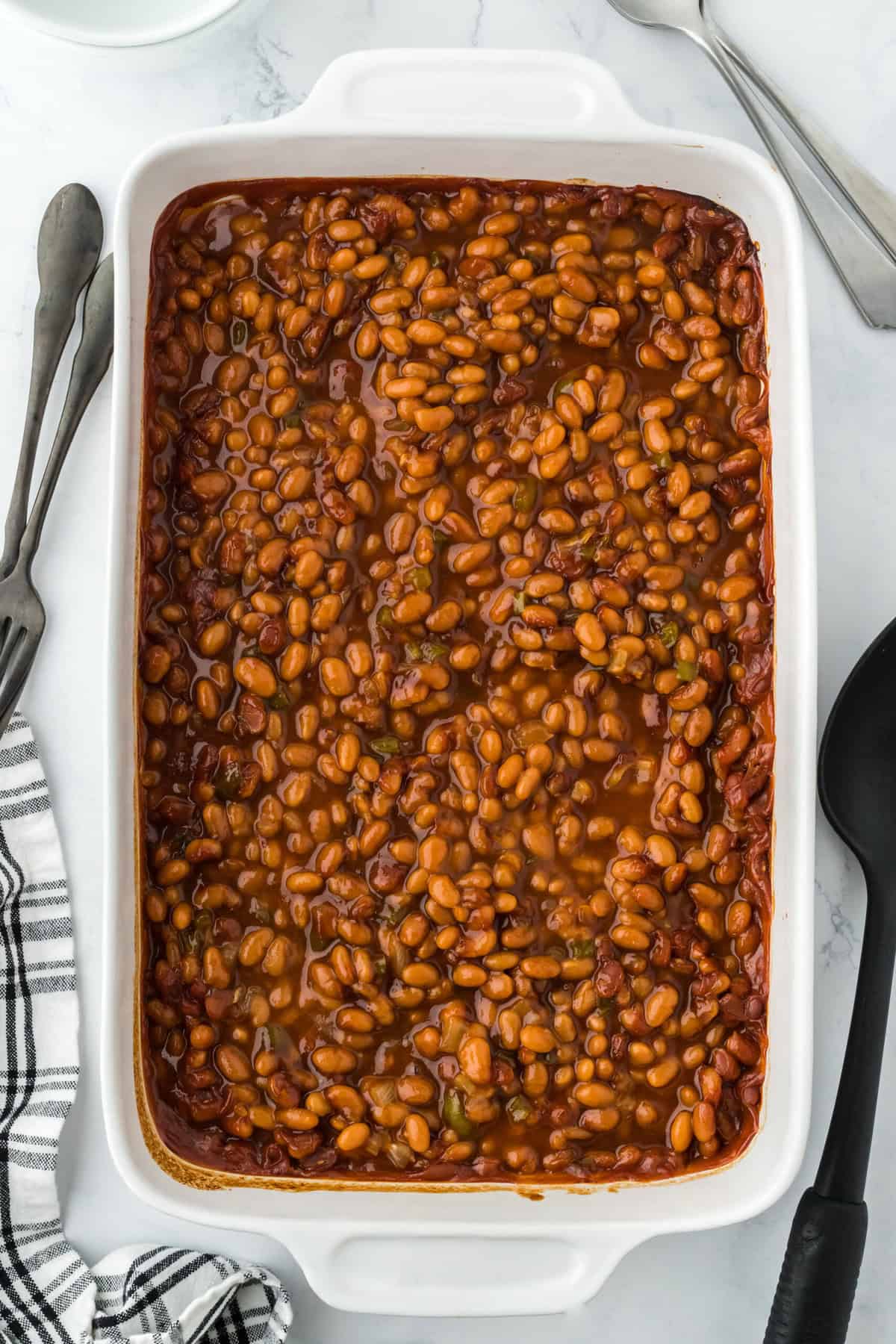 A large dish of homemade baked beans recipe in a white casserole dish on a white background with a black spoon on the side
