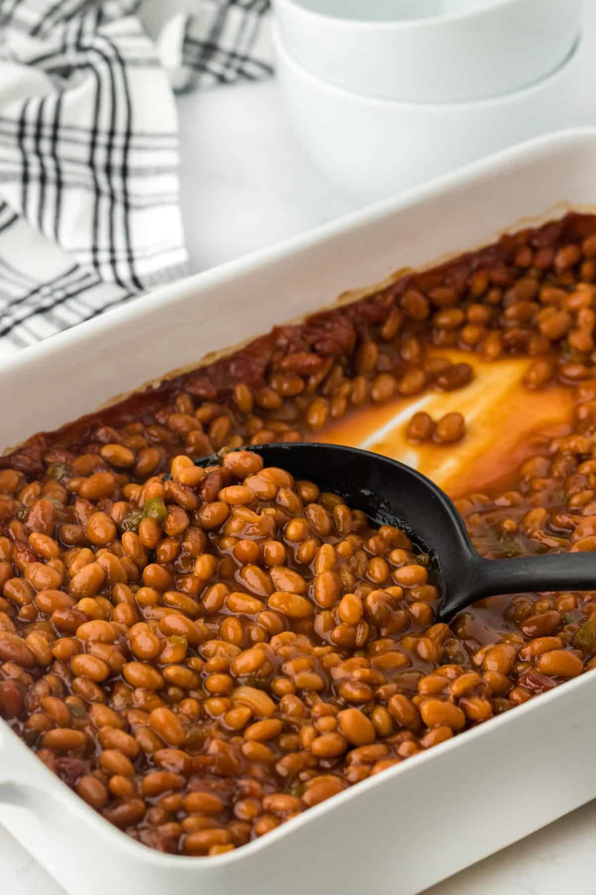 A large white dish of easy baked beans on a white background with a black spoon scooping it up with black and white striped napkin