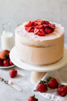 A strawberry buttercream frosted layer cake with strawberry slices on top on a cake plate with berries near by