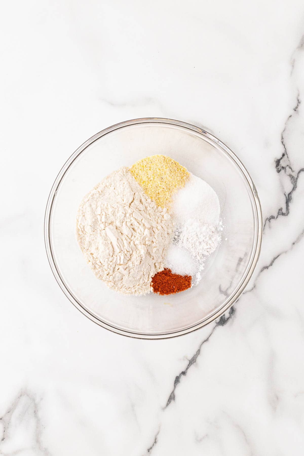 Flour, cornmeal, sugar, baking powder, salt, and cayenne pepper in a glass bowl on a white marble surface