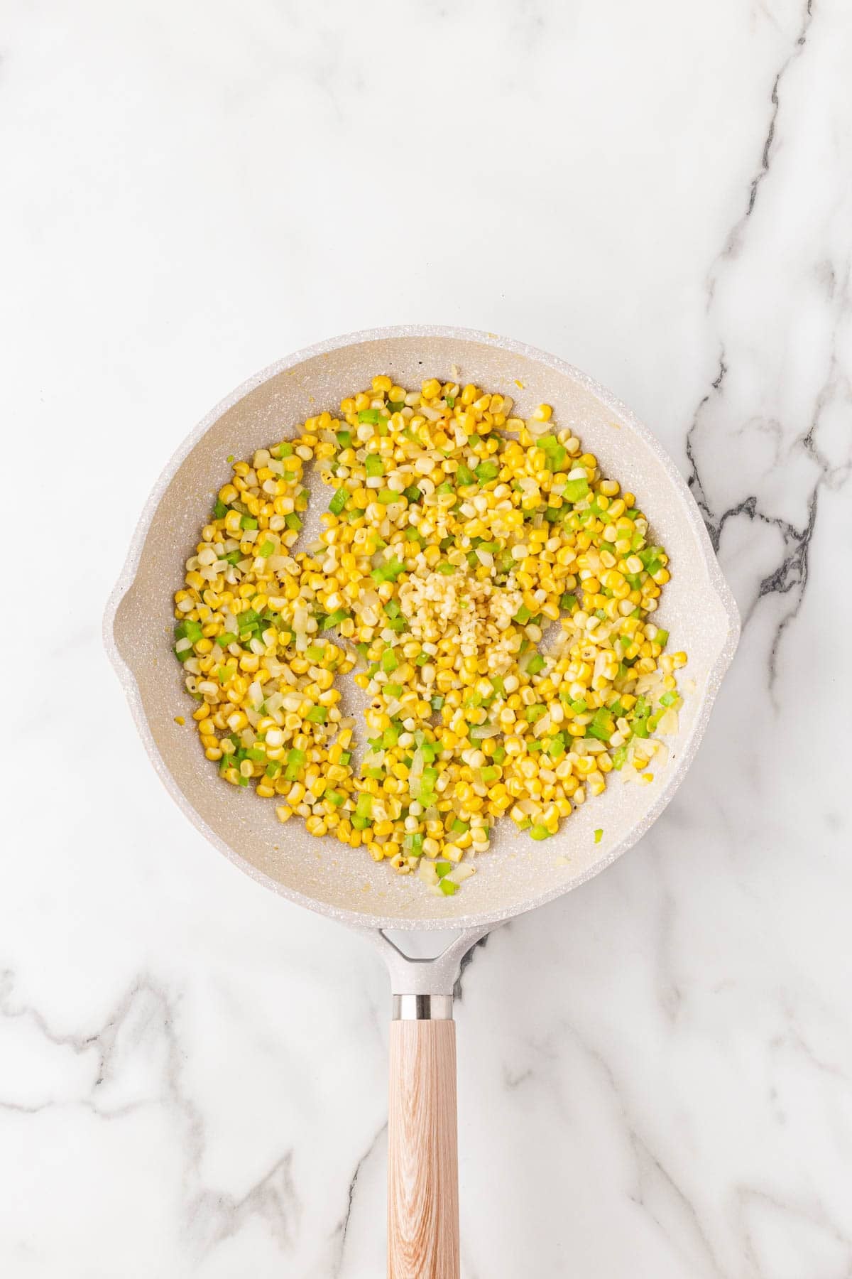Corn, onion, green pepper, and garlic sautéing in a skillet on a white marble surface