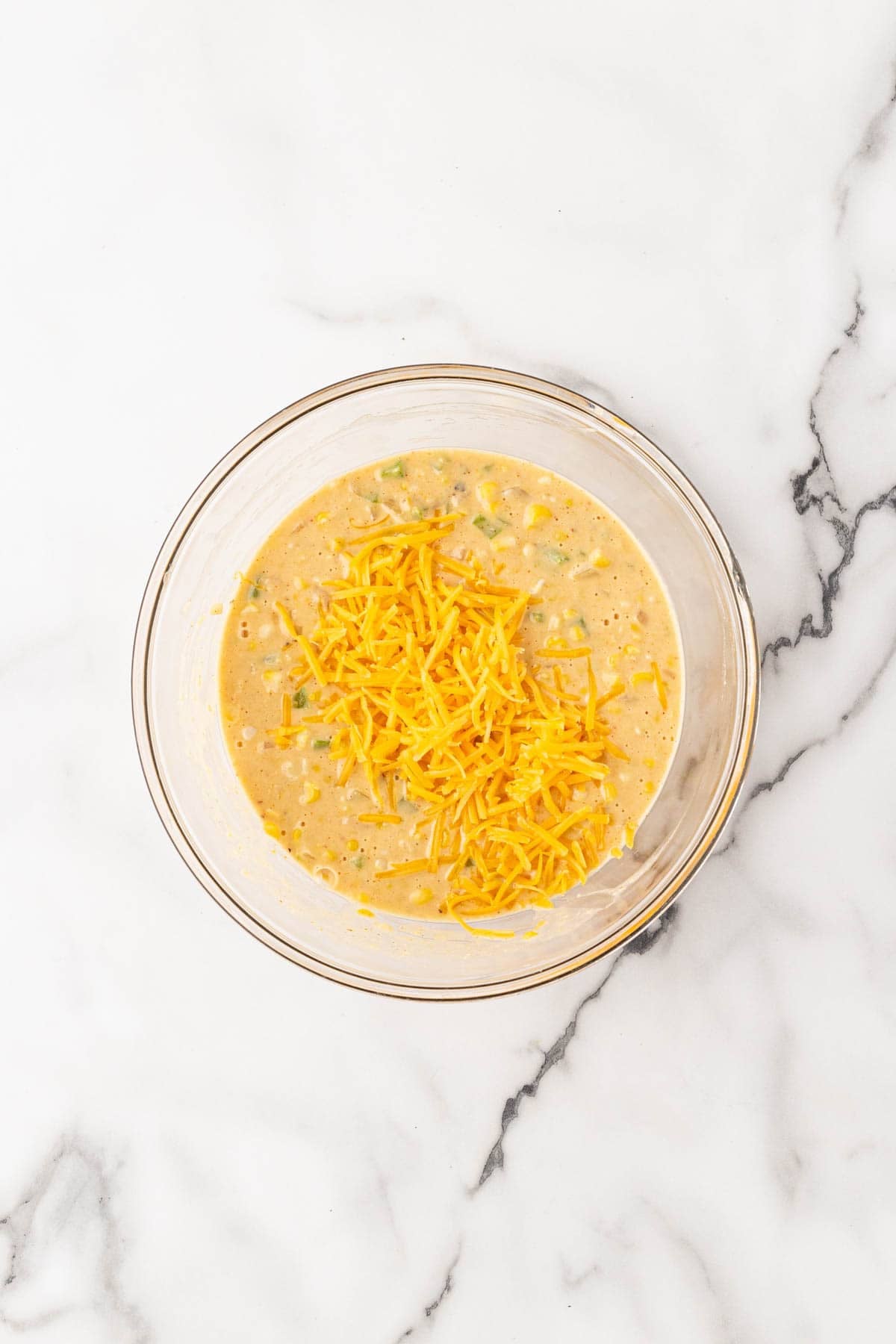 Shredded cheddar cheese and veggie mixture added to the batter in a glass bowl on a white marble surface