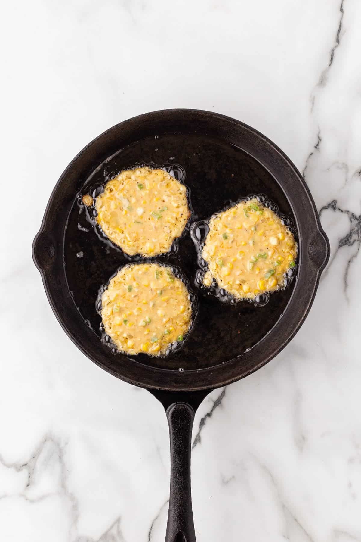 Corn fritter batter frying in a cast iron skillet on a white marble surface