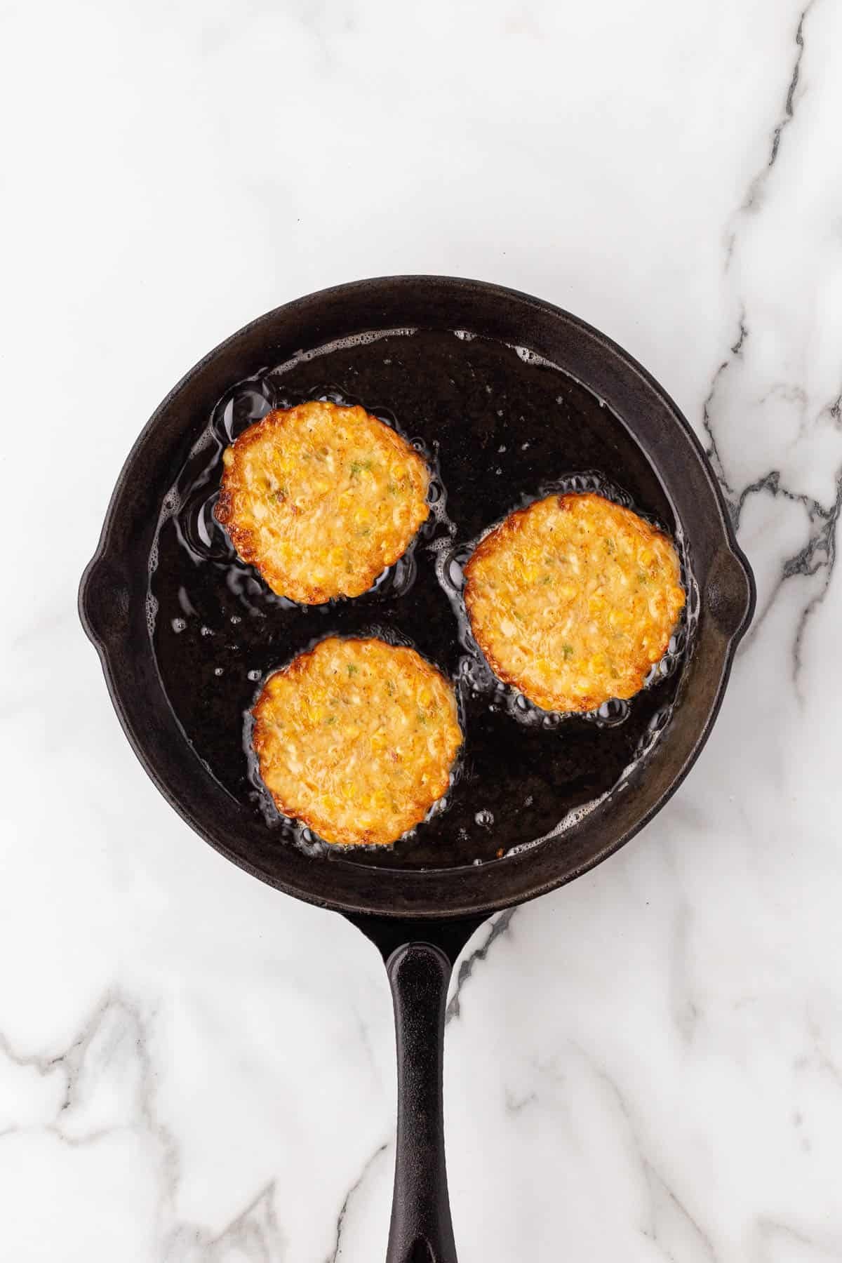 Golden brown corn fritters frying in a cast iron skillet on a white marble surface