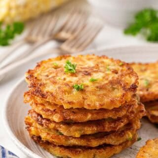 Stack of golden corn fritters on a ceramic plate with corn on the cob in the background