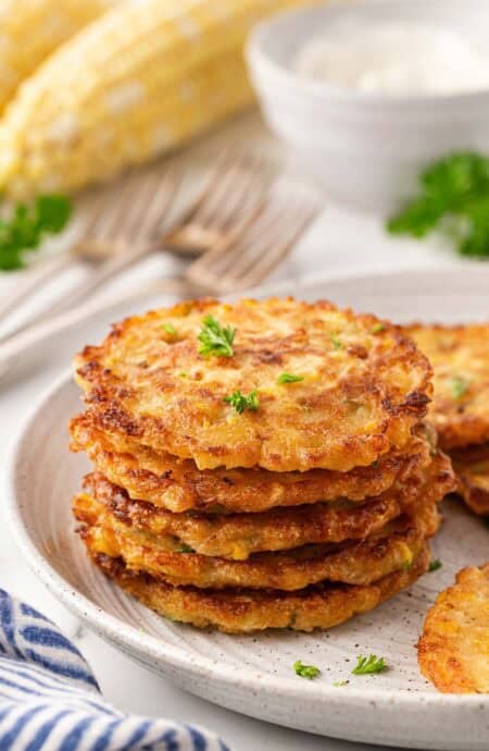 Stack of golden corn fritters on a ceramic plate with corn on the cob in the background
