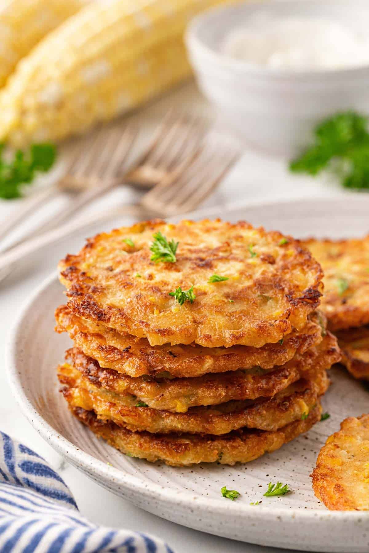 Stack of golden corn fritters on a ceramic plate with corn on the cob in the background