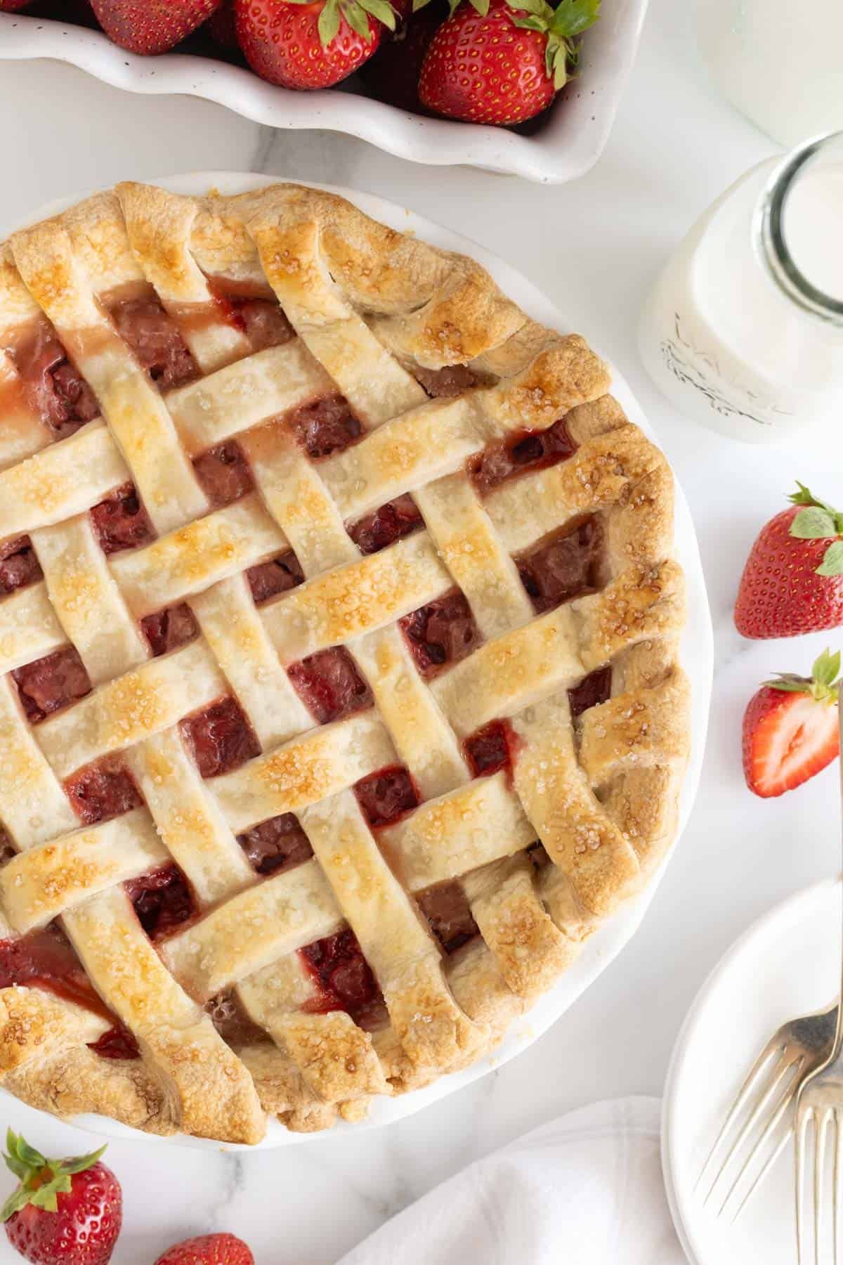 Overhead shot of a golden-brown strawberry pie with a lattice crust, surrounded by fresh strawberries and a glass of milk