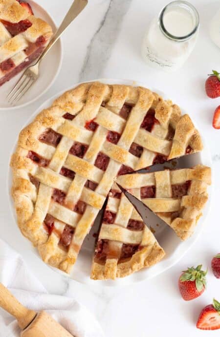 Sliced strawberry pie with one slice removed, surrounded by fresh strawberries, milk, and more pie