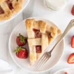 Slice of strawberry pie served on a white plate with two whole strawberries and a fork, surrounded by fresh strawberries and a glass of milk