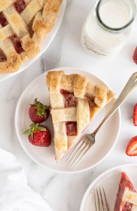 Slice of strawberry pie served on a white plate with two whole strawberries and a fork, surrounded by fresh strawberries and a glass of milk