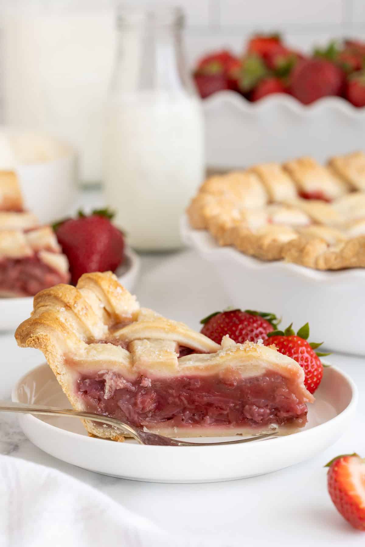 Closeup of a slice of strawberry pie on a plate with fresh strawberries and a fork on the side, and milk in the background