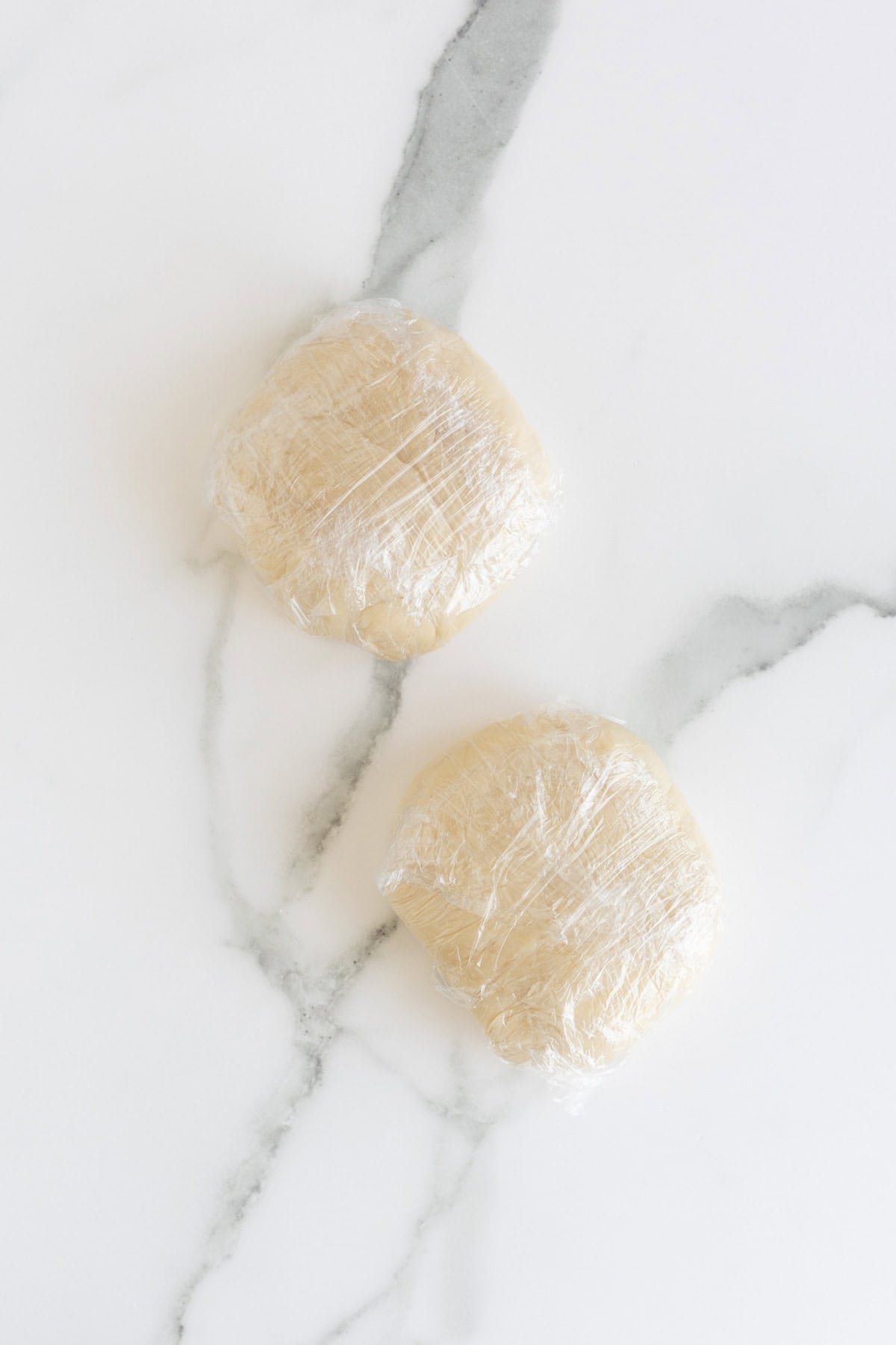Two dough discs wrapped in plastic wrap on marble counter