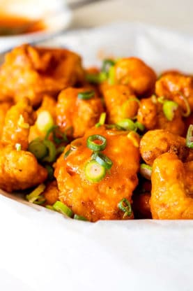 A close up of a pile of buffalo cauliflower with green onions on top for pops of color against a white background