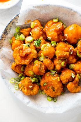 A pile of delicious golden fried cauliflower tossed in buffalo sauce and topped with green onions against white parchment paper