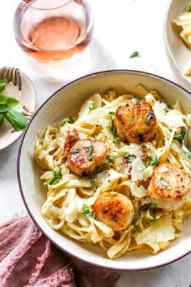 A bowl of creamy garlic pasta with scallops next to a glass of rose' against a white background