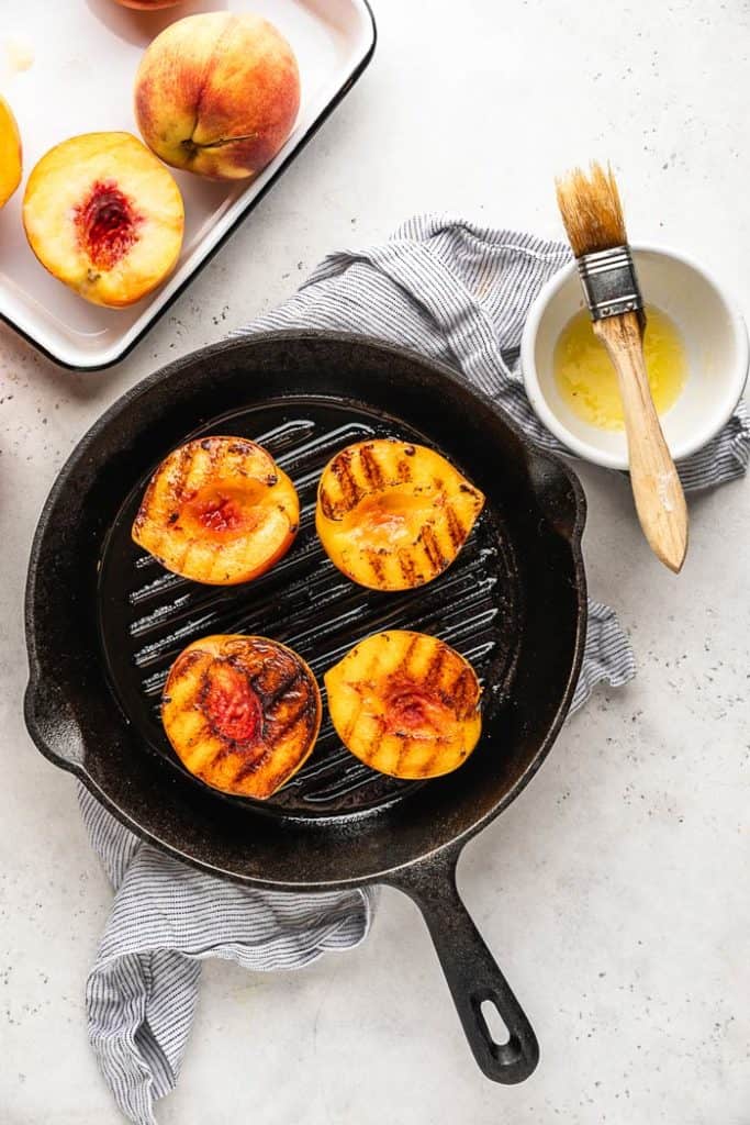 Peach halves on a cast iron grill pan with grill marks and a butter bowl with a brush