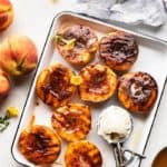 A white tray of brown sugar grilled peach halves with a scoop of vanilla ice cream next to fresh peaches
