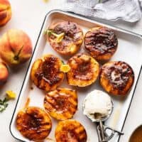 A white tray of brown sugar grilled peach halves with a scoop of vanilla ice cream next to fresh peaches