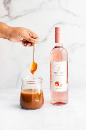 A spoon dipped into a glass jar filled with spiked caramel sauce next to a bottle of Barefoot Fruitscato 