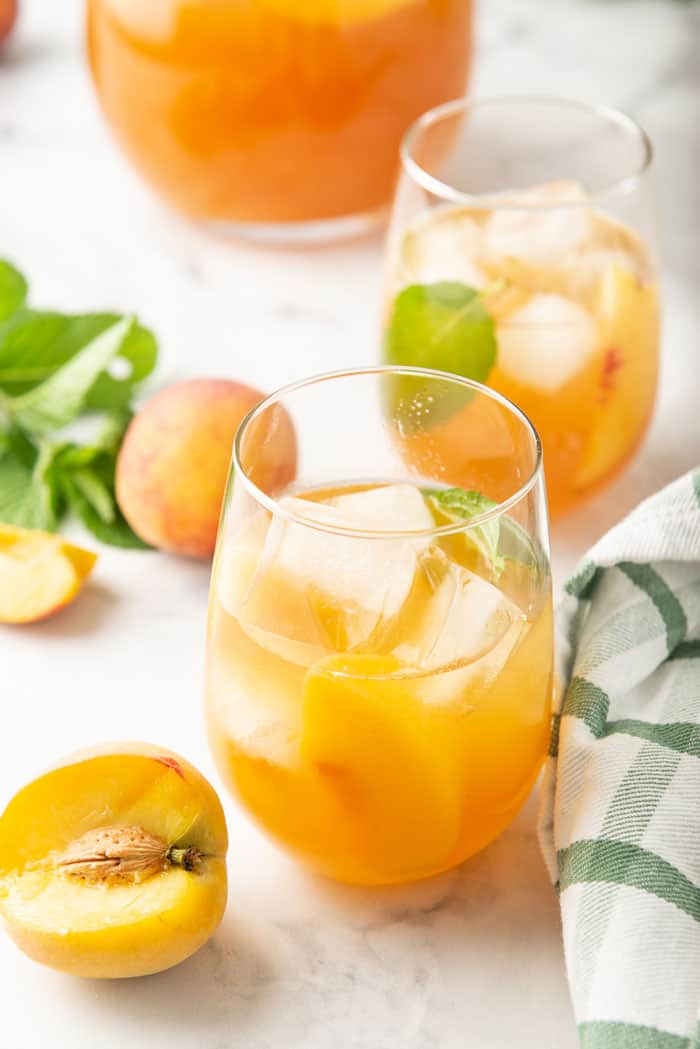 Peach sweet tea in two glasses surrounded by a pitcher, fresh peaches and mint