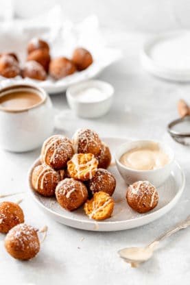 A white plate with pumpkin fritters powdered with sugar and drizzled with glaze next to a cup of coffee and bowl of glaze