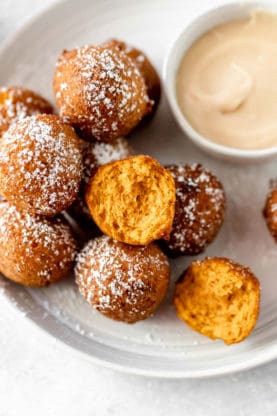A pile of pumpkin fritters with powdered sugar on top next to a cream cheese glaze bowl