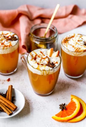Three mugs of spiced cider with whipped cream and caramel sauce surrounded by a coral napkin