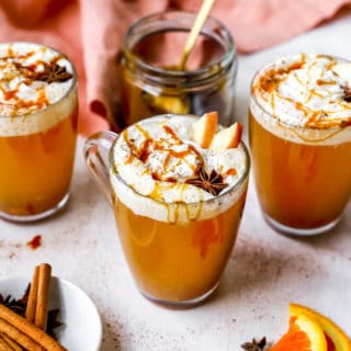 Three mugs of spiced cider with whipped cream and caramel sauce surrounded by a coral napkin